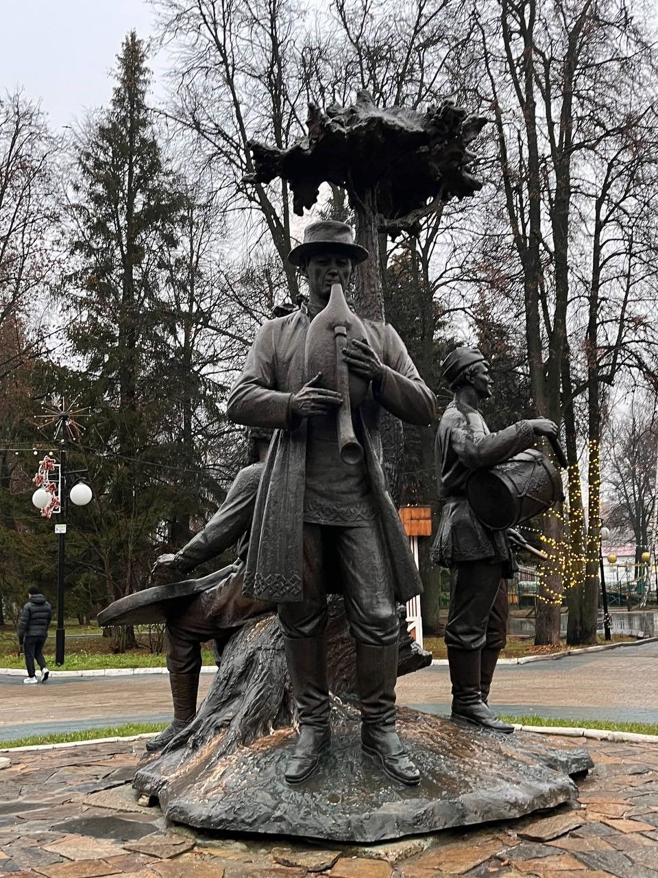 На фотографии изображен памятник &laquo;Древо жизни&raquo; г. Йошкар-Ола. Скульптура состоит из трёх фигур &ndash; старца, мужчины и мальчика, &ndash; играющих на национальных инструментах: гуслях, волынке и барабане.